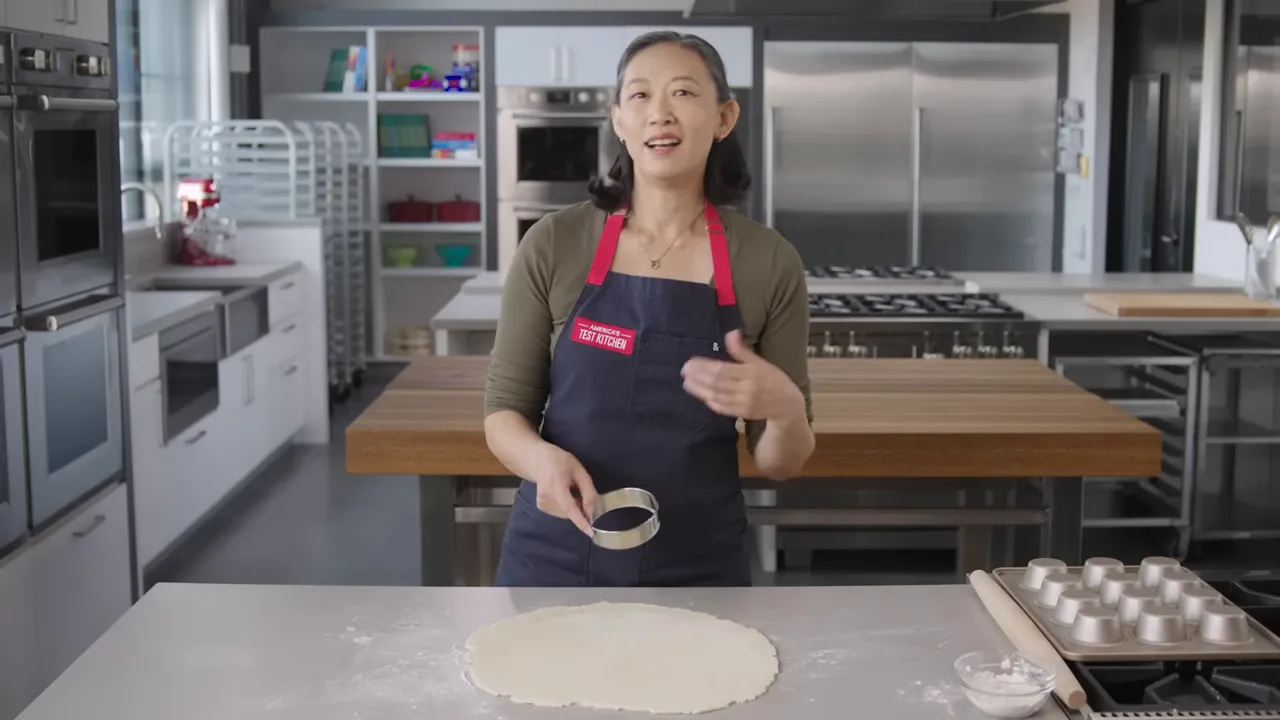 Rolled pastry dough on a countertop with a cook holding a round cutter and mini tart pans nearby.