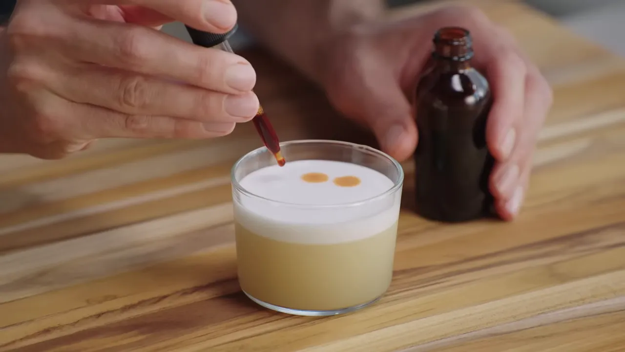 dropper placing bitters dots on a glass of cocktail with egg-white foam