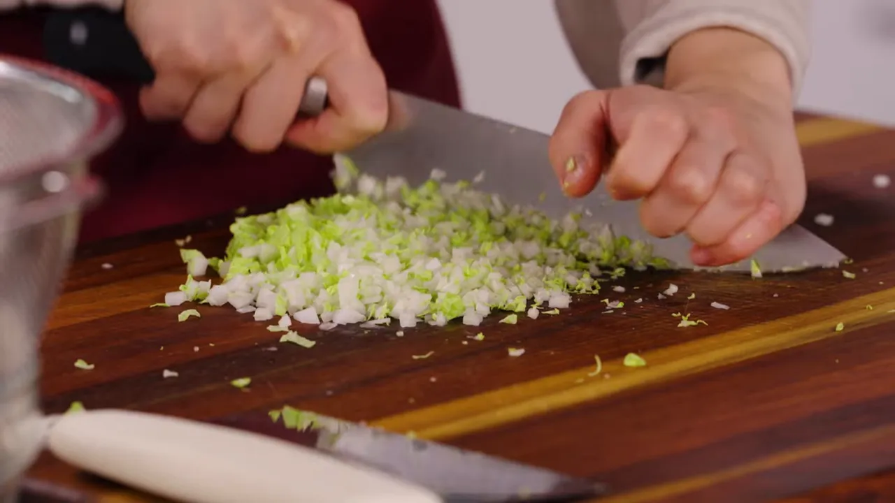 Pile of finely minced Napa cabbage on a wooden cutting board with a cleaver and hands visible.