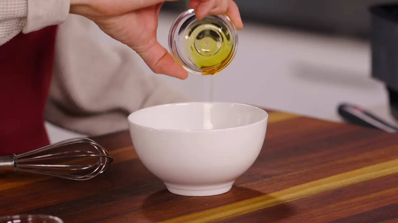 Hand pouring a small glass bowl of oil into a white mixing bowl on a wooden board