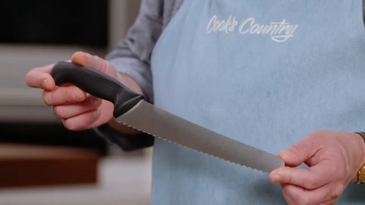 Presenter holding a 10‑inch serrated bread knife showing the full blade and handle against a Cook's Country apron.