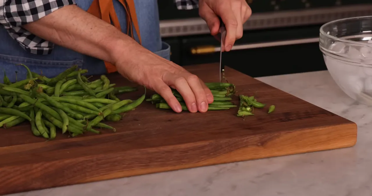 Hands trimming the ends off fresh green beans on a wooden cutting board with an ice bowl visible to the side