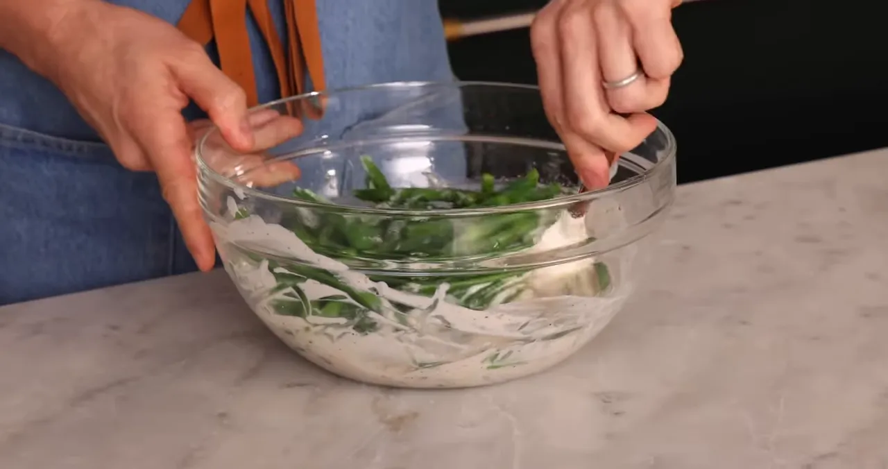 Hands tossing blanched green beans in a creamy mushroom sauce in a glass bowl
