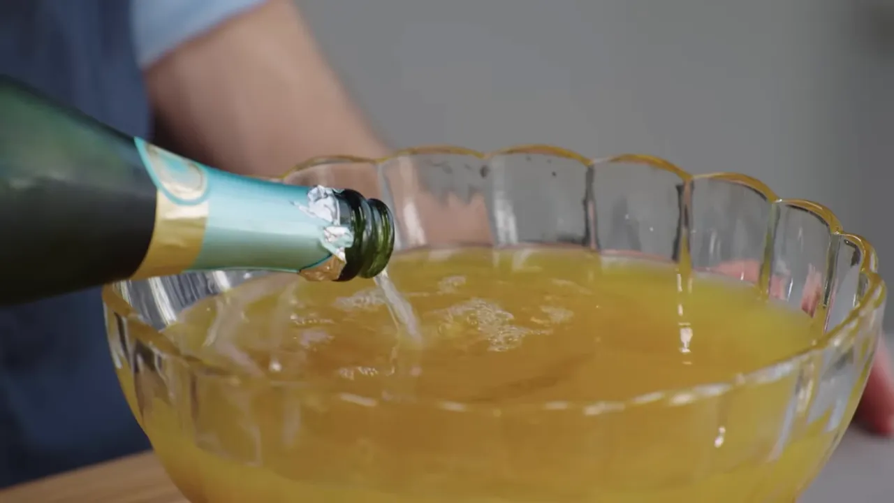 Close-up of a sparkling wine bottle pouring a stream into a glass punch bowl filled with orange punch