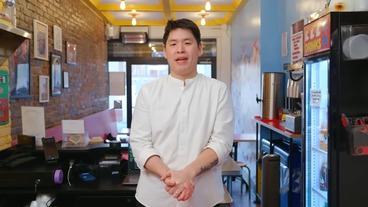 Chef standing and talking in a bright sandwich shop with prep counter and refrigerated display visible