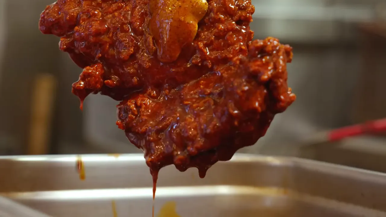 Close-up of a fried chicken thigh coated in red chili oil, dripping into a stainless pan