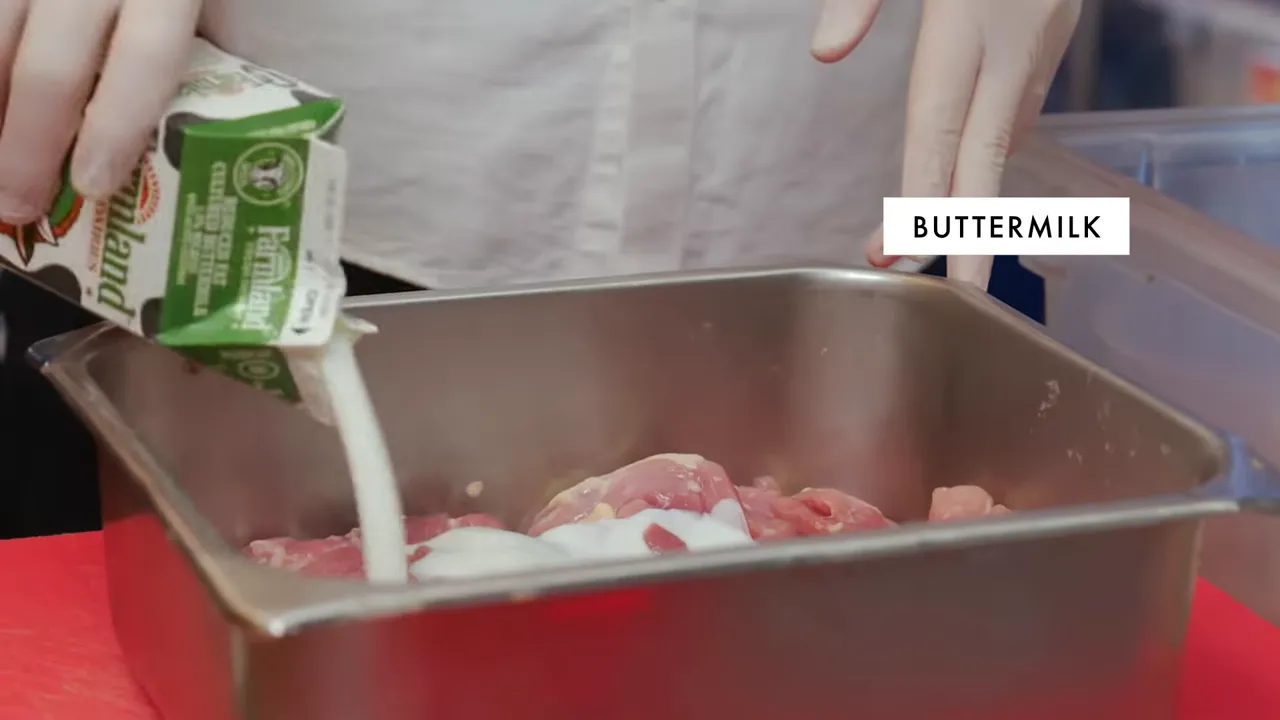 Buttermilk being poured from a carton over chicken thighs in a metal pan