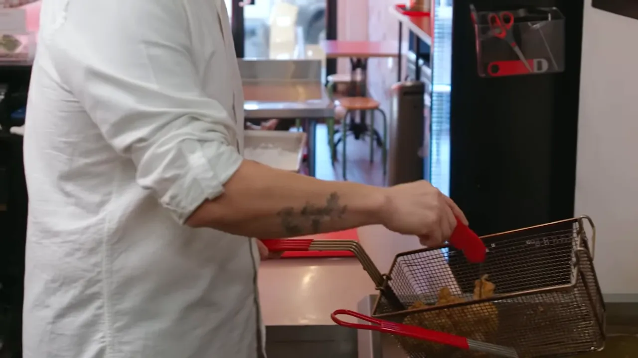 Hand removing a basket of fried chicken pieces from a commercial fryer using a red silicone-tipped tool