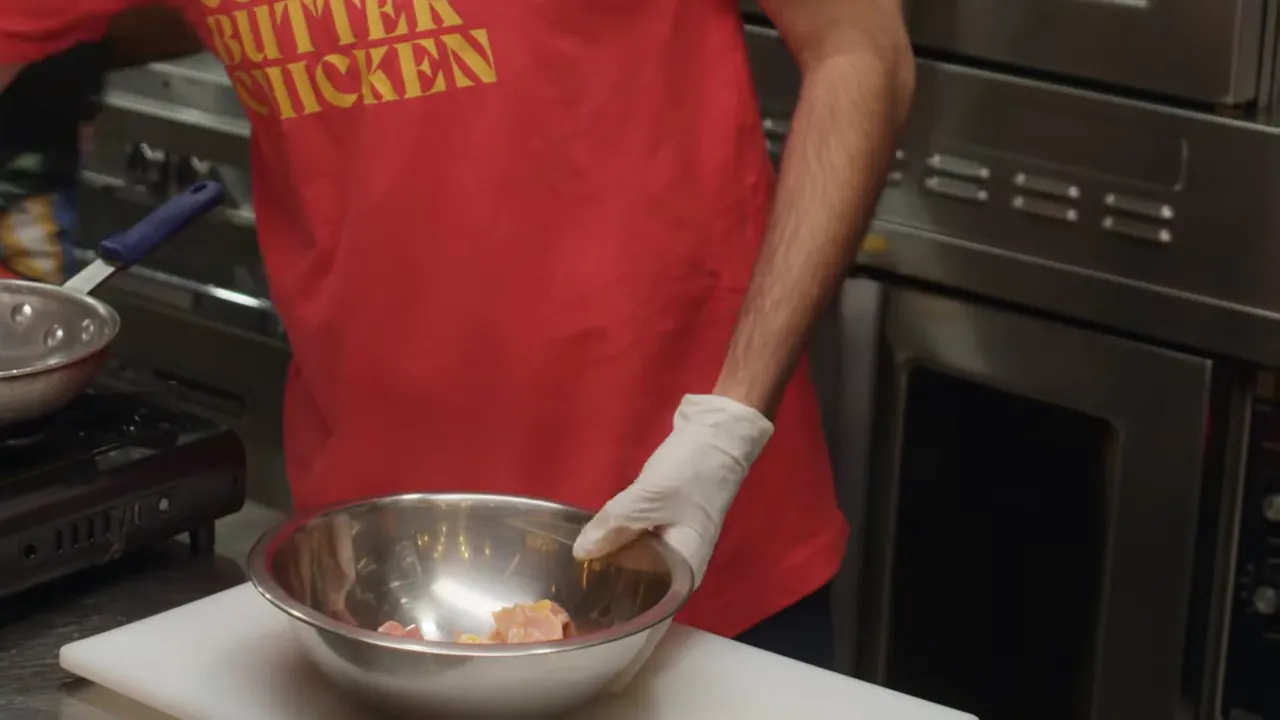 Gloved hand holding a stainless steel bowl with cubed chicken on a cutting board ready to be seasoned