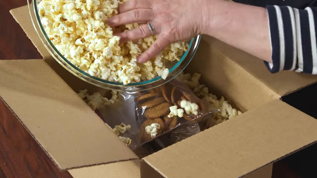 hand pouring a bowl of popcorn into a cardboard box to cushion a zipper bag of cookies placed in the center