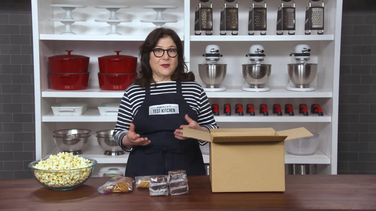 Presenter with an open shipping box on a table and assorted packaged cookies and plastic-wrapped bars arranged in front.