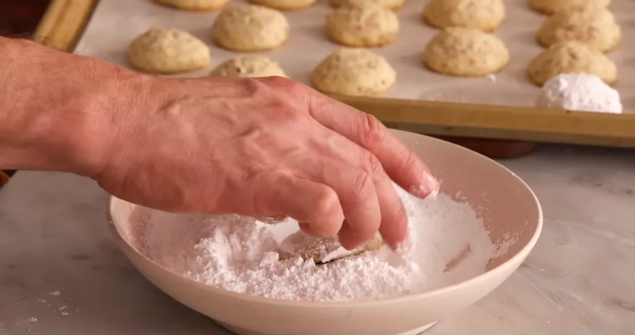Close-up of a hand rolling a baked cookie in powdered sugar with a tray of cookies in the background