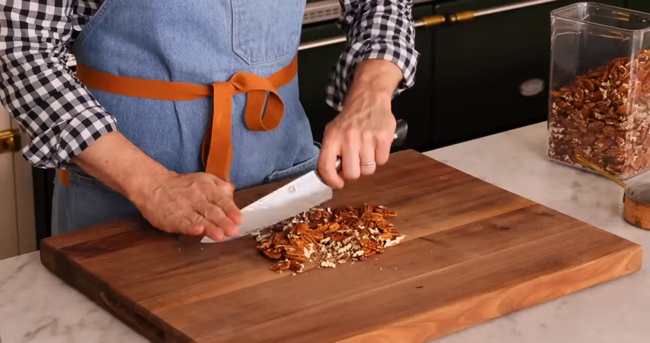 Close-up of hands chopping pecans on a wooden board with a container of pecans nearby
