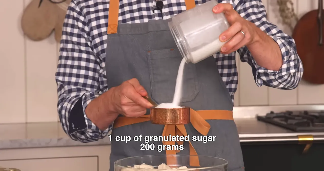 Close-up of granulated sugar being poured into a measuring cup with on-screen text '1 cup of granulated sugar 200 grams' above a bowl of biscuit pieces