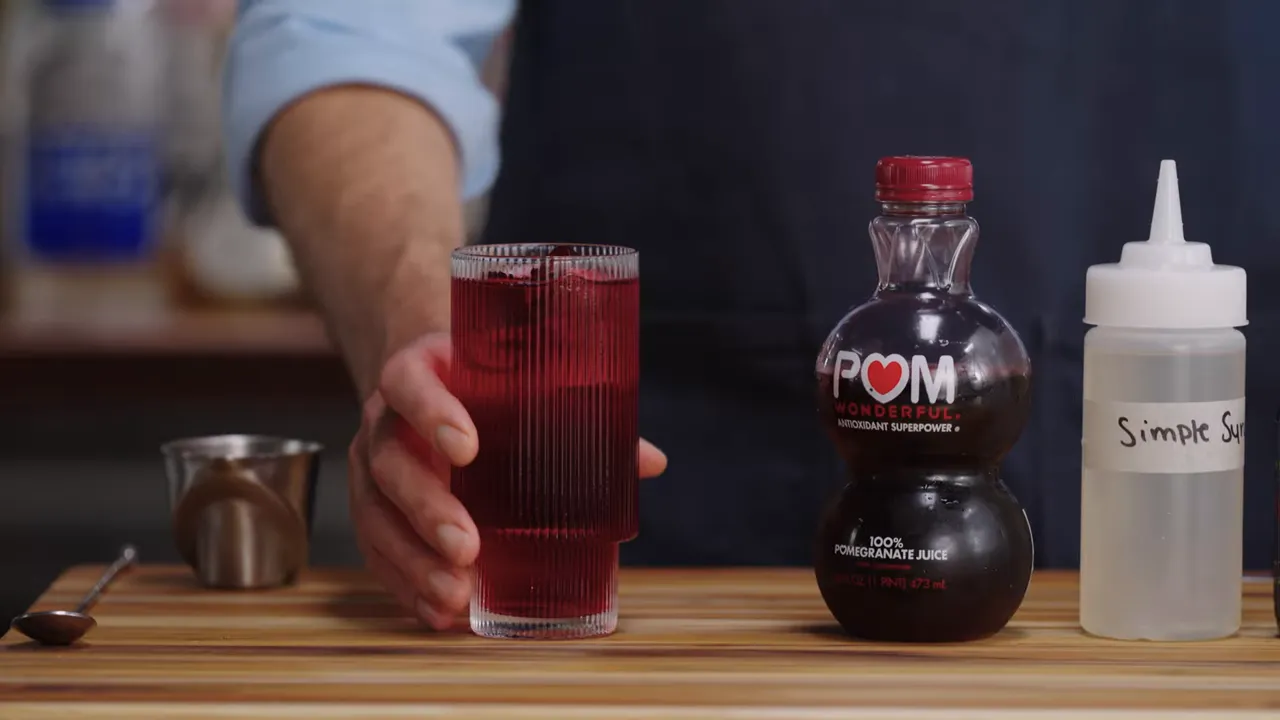 Ribbed highball glass with deep-red pomegranate spritzer, POM pomegranate juice bottle and a squeeze bottle labeled simple syrup on a wooden board.
