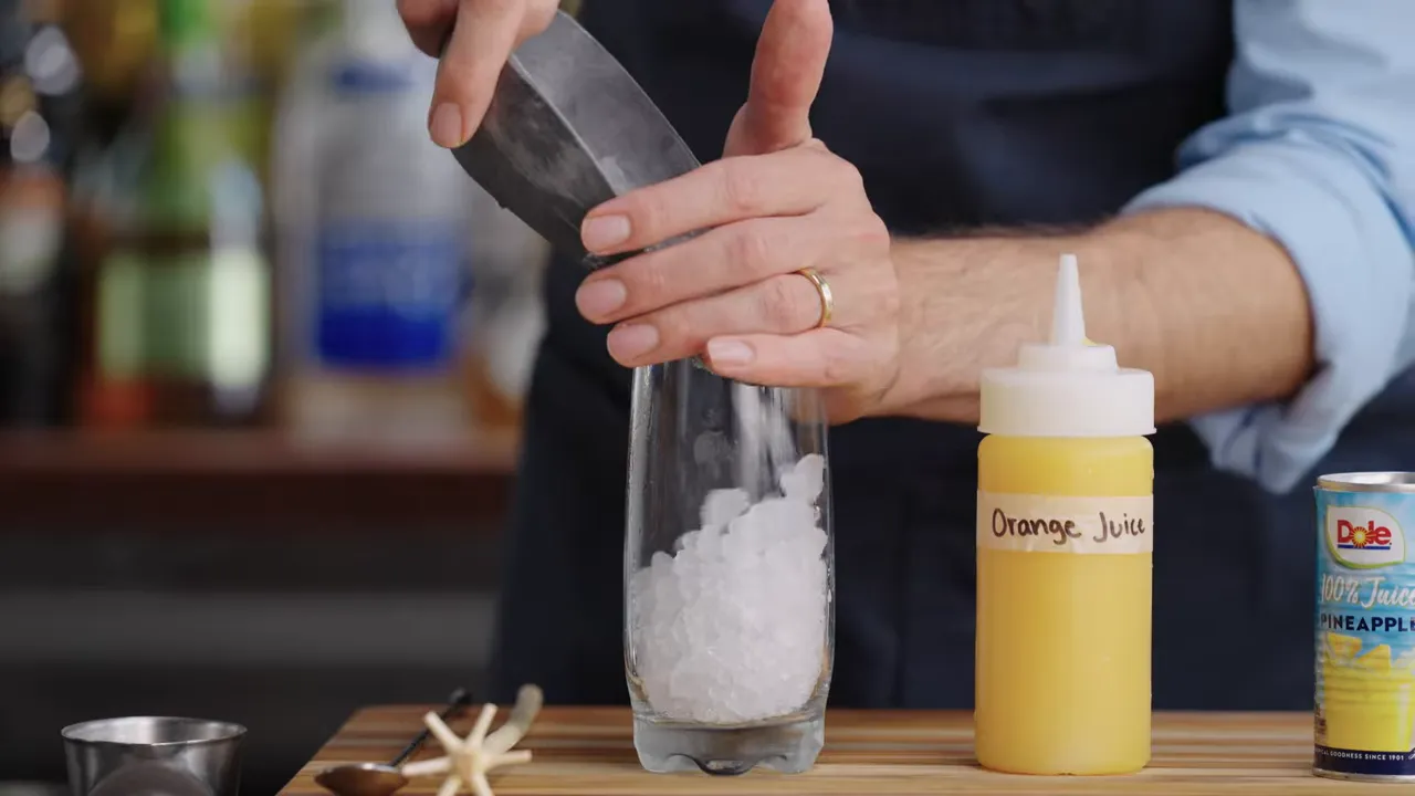 hands packing crushed ice into a tall glass with a labeled squeeze bottle of orange juice and a can of pineapple juice visible