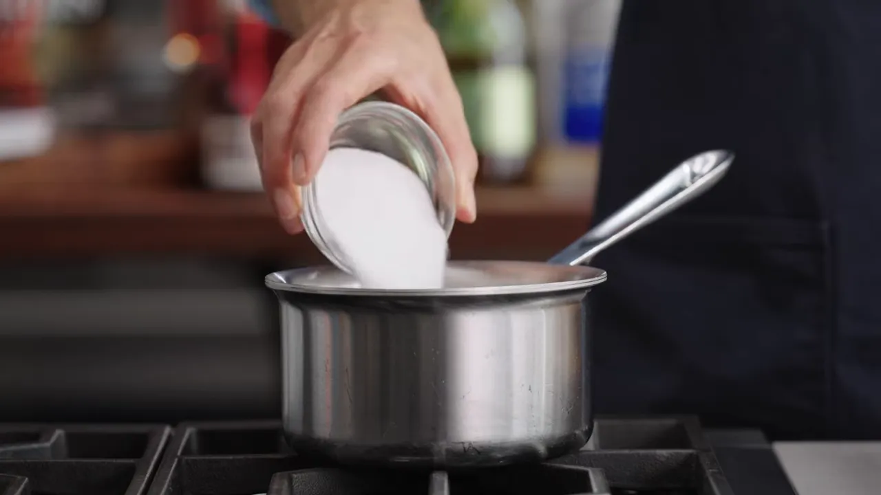 hand pouring sugar from a small bowl into a stainless steel saucepan on a stove