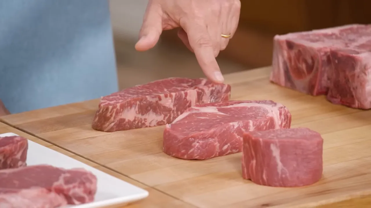 Hand pointing to a selection of raw steaks (tenderloin medallion, strip and ribeye) on a cutting board