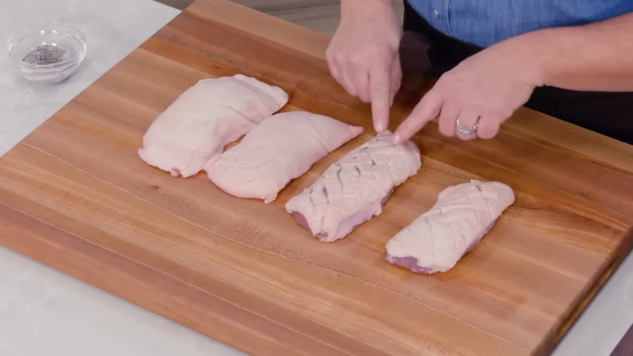 Raw duck breasts on a wooden cutting board with fat scored in a hatch pattern and hands pointing to the cuts