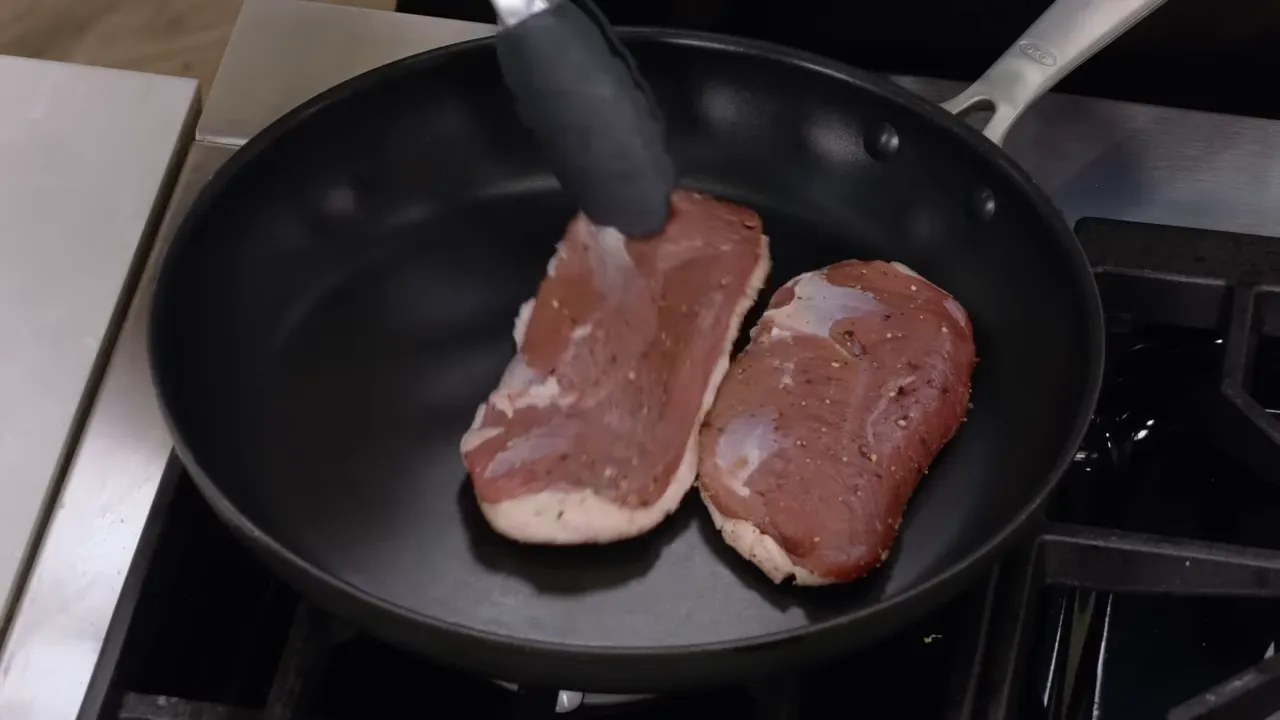 Two duck breasts being placed in a skillet with tongs, fat side down