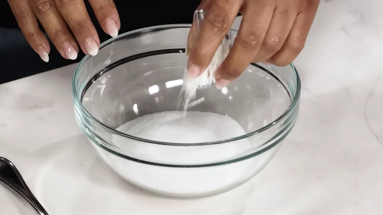 Hand sprinkling a small amount of cornstarch into a glass bowl of granulated sugar.