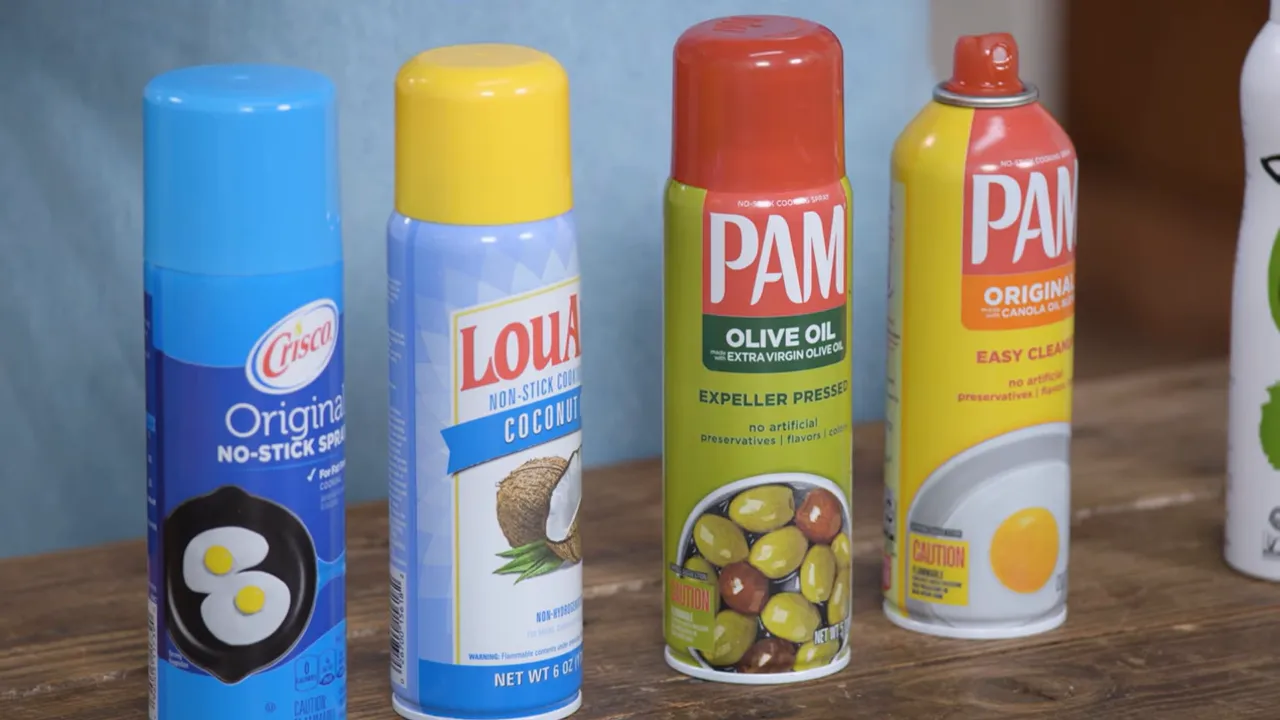 Four cooking spray cans (blue, yellow, green and orange-labeled) lined up on a wooden counter, labels and caps visible.
