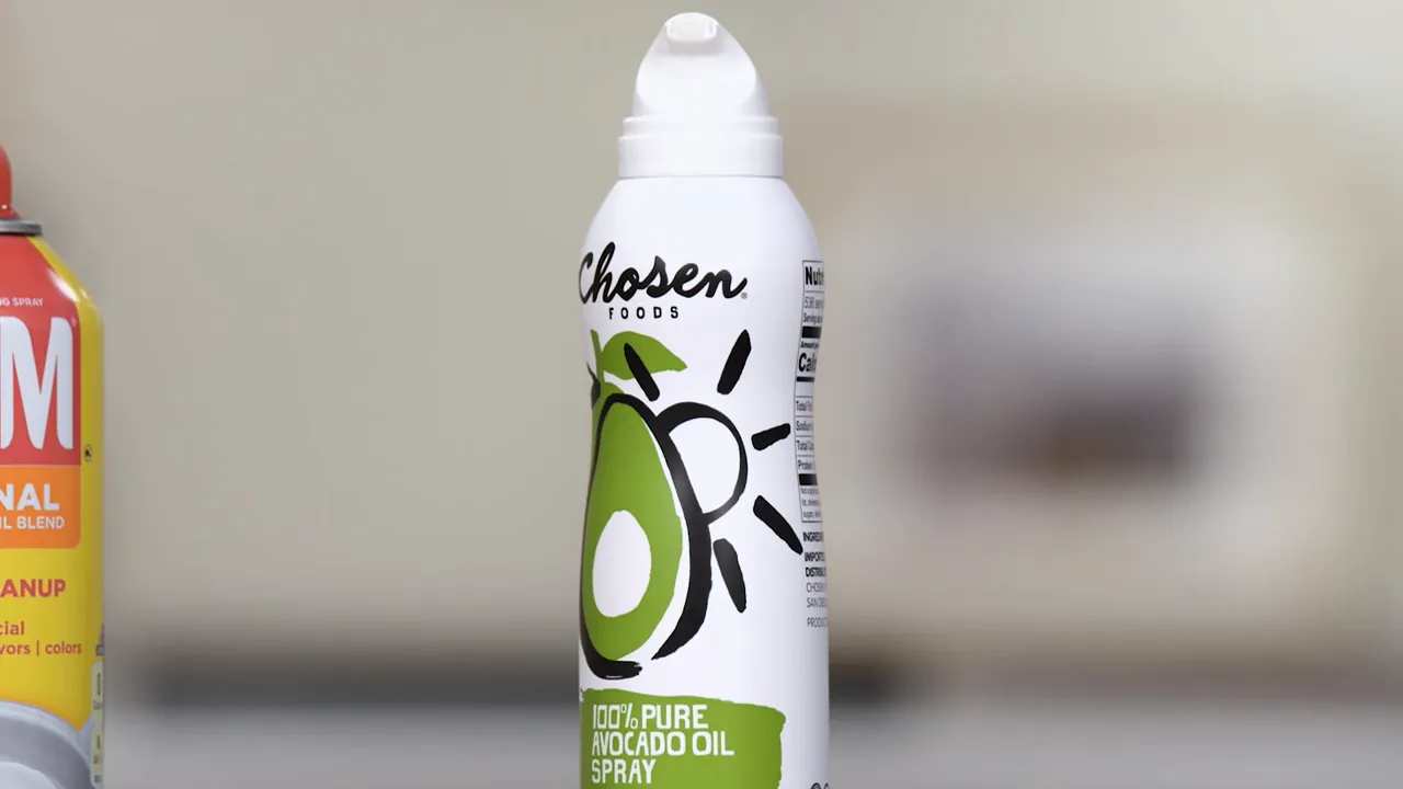 Centered close-up of a white 'Chosen' avocado oil spray can showing the top nozzle and green avocado artwork against a blurred kitchen background.