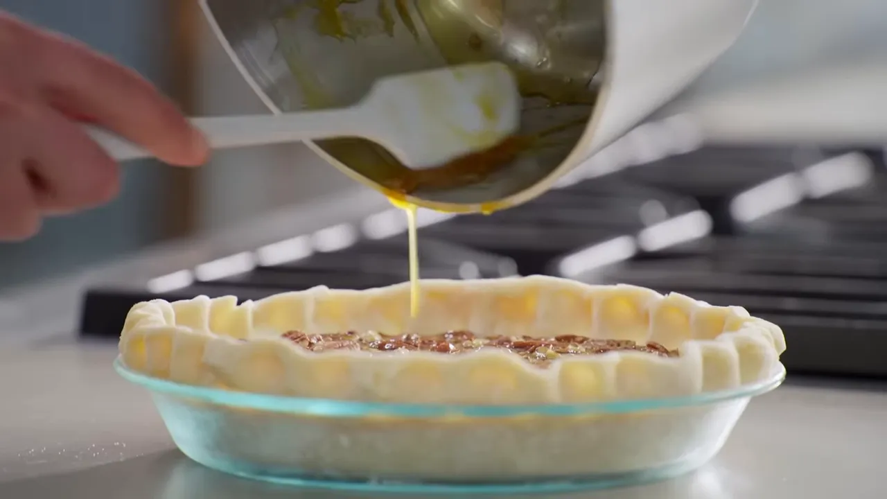 Custard being poured from a saucepan into a pecan-filled pie shell with a spatula