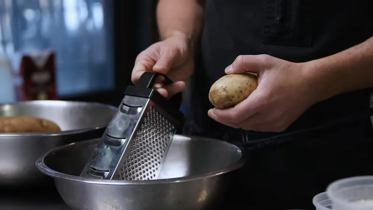 close-up of hands holding an Idaho potato and a box grater over a metal mixing bowl