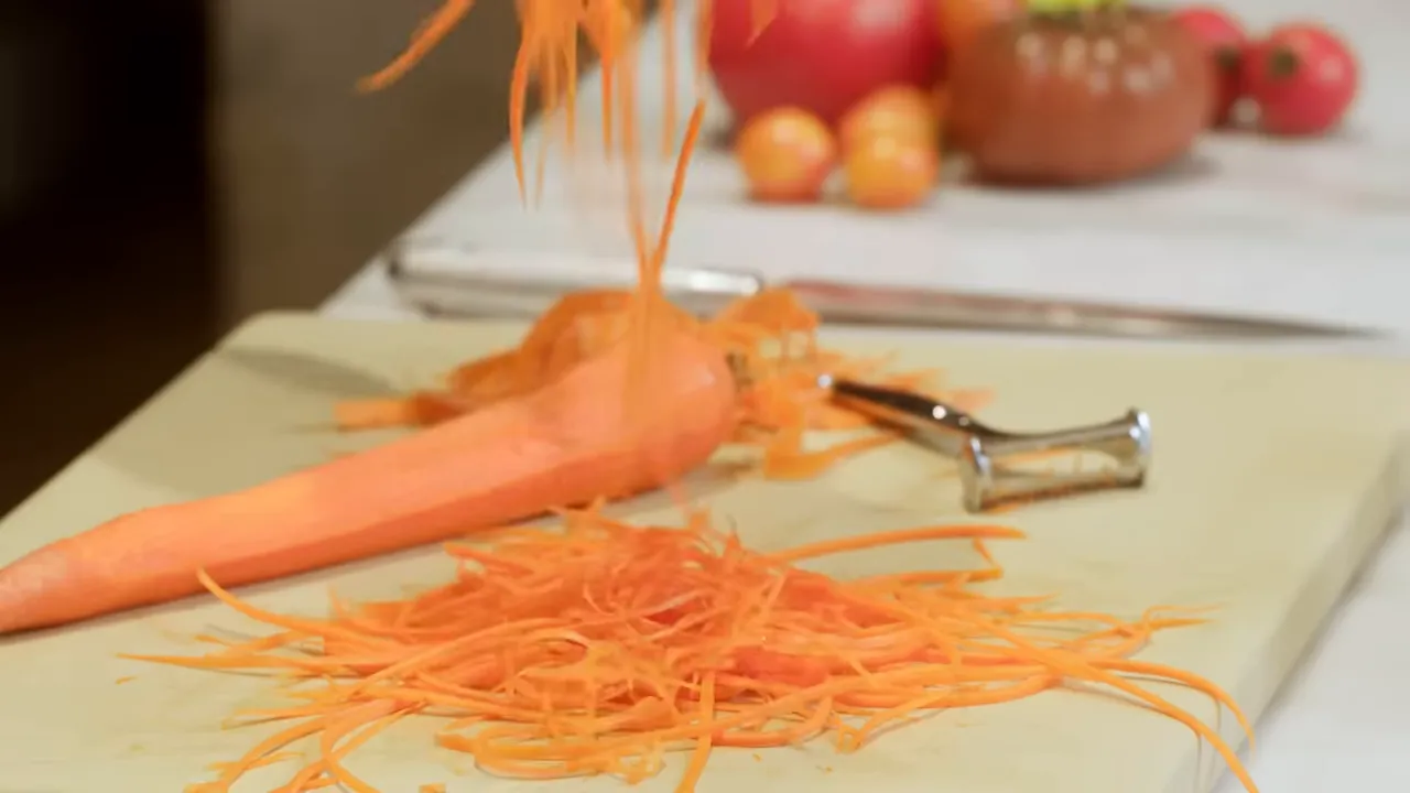 pile of thin julienne carrot ribbons on a cutting board with a julienne peeler and peeled carrot