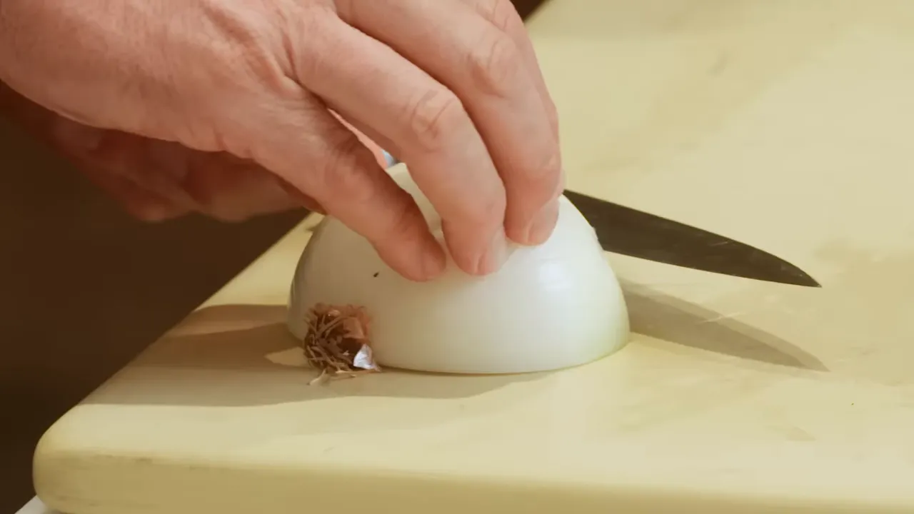 Hand stabilizing an onion half with the root intact as a knife slices on a cutting board