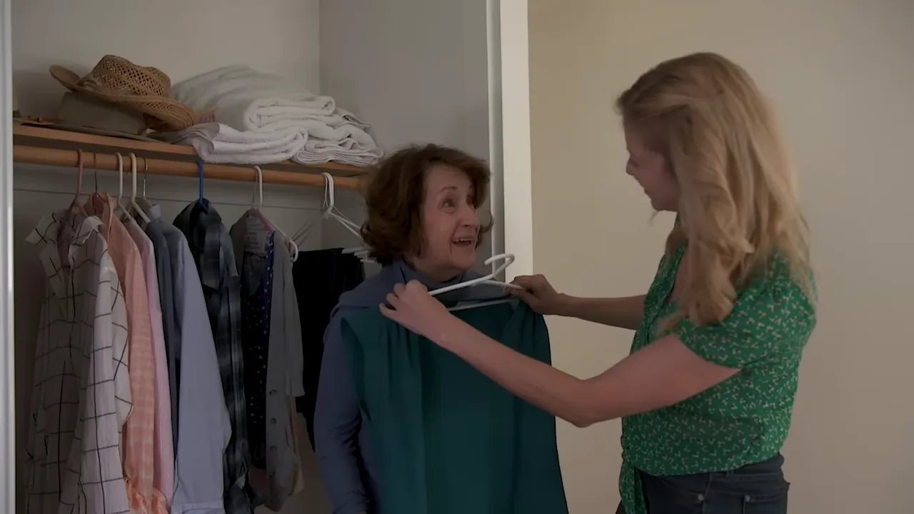 caregiver holding a green top in front of an older woman near an open closet