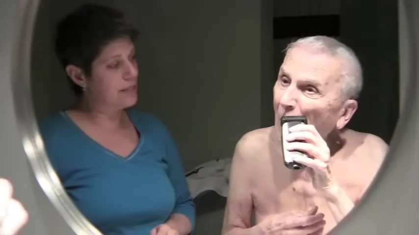 Older adult using an electric shaver while another person stands nearby in a bathroom mirror view