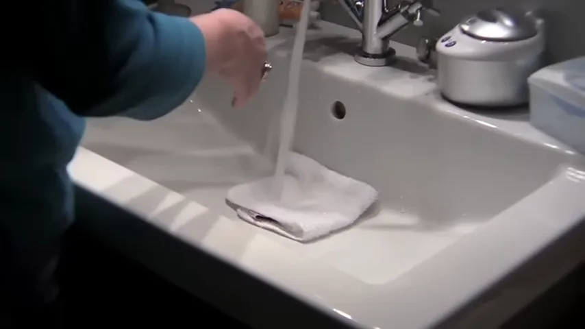 Hands rinsing a washcloth in a bathroom sink under running water