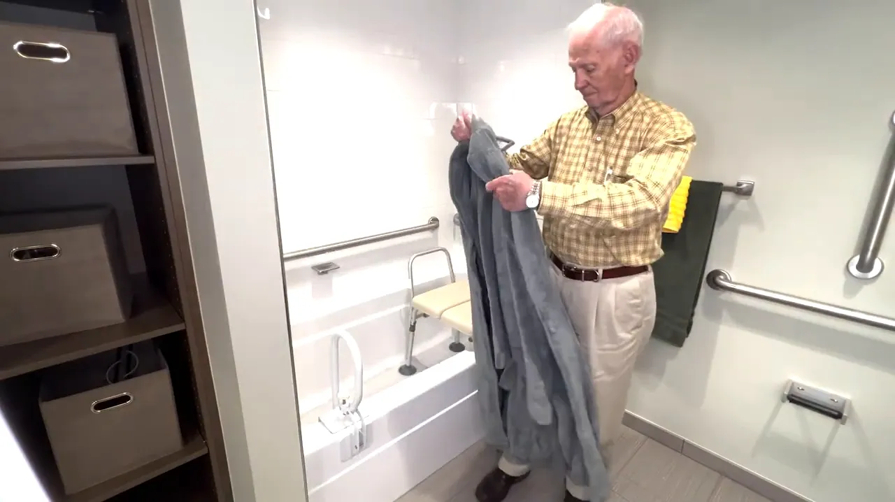 Older adult holding a gray bathrobe beside the bathtub