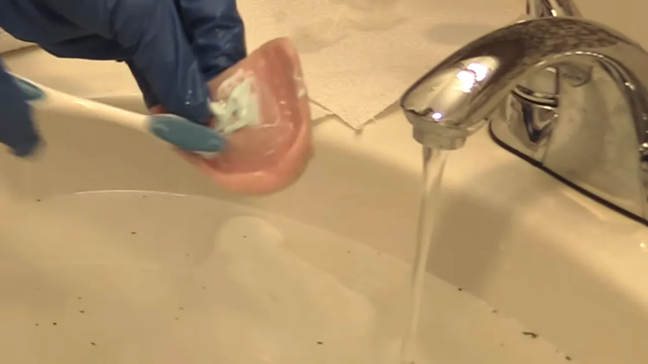 Gloved hands brushing the inside surface of an upper denture near a running faucet