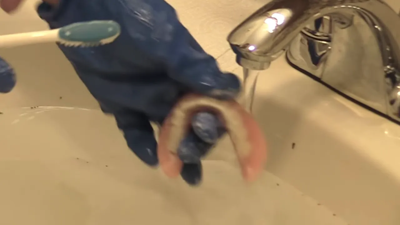 Gloved hands holding a lower denture under running water while brushing it