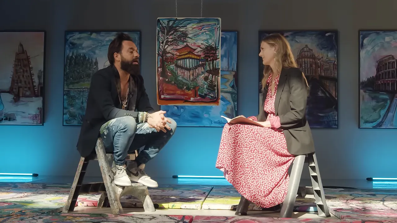 Two people seated in studio facing each other with a central hanging painting behind them