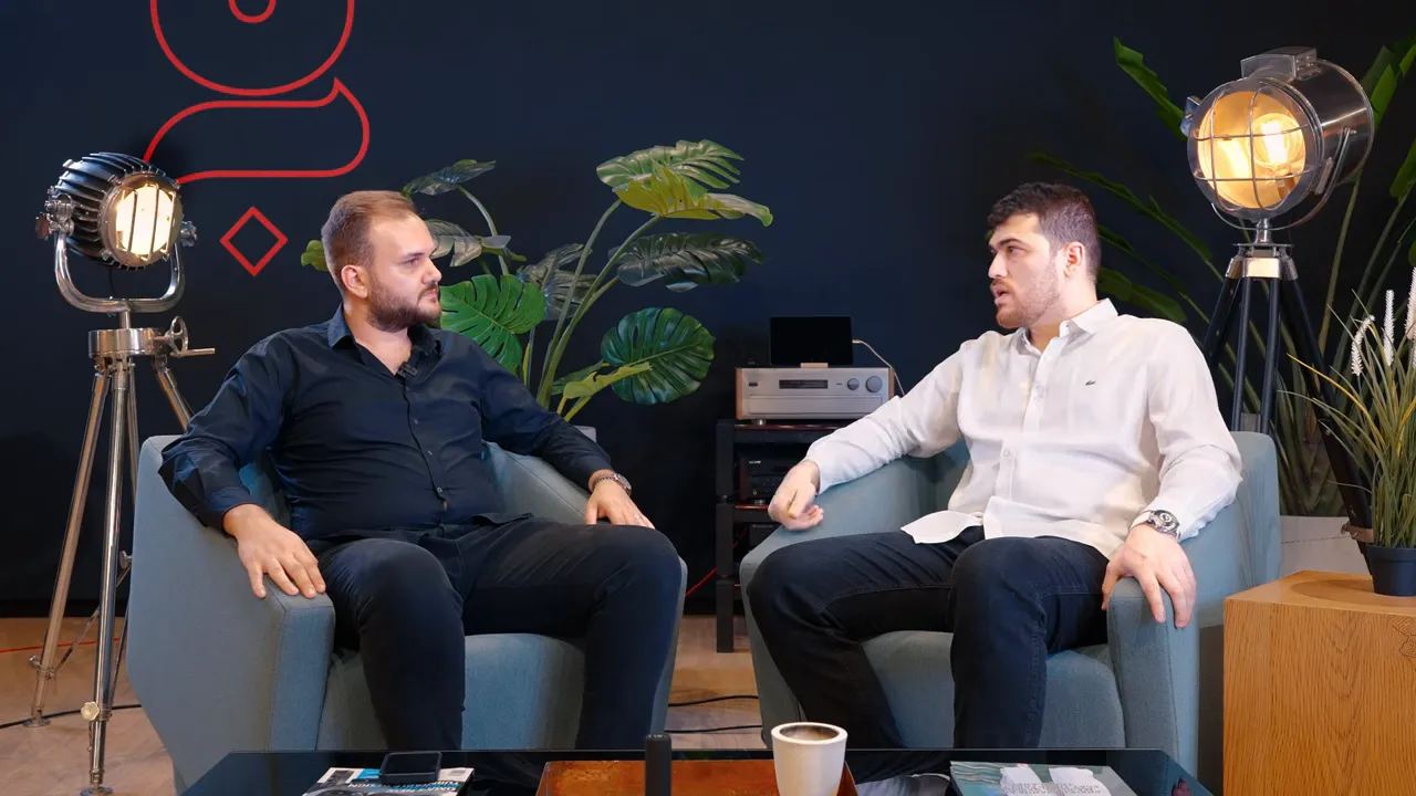 İki kişi koltuklarda sohbet ediyor; sağdaki kişi el hareketiyle fikir belirtiyor, g&ouml;r&uuml;nt&uuml; net ve dengeli aydınlatılmış.