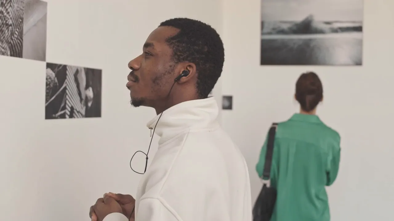 Two gallery visitors viewing framed photographs on a white wall, one listening to an audio guide