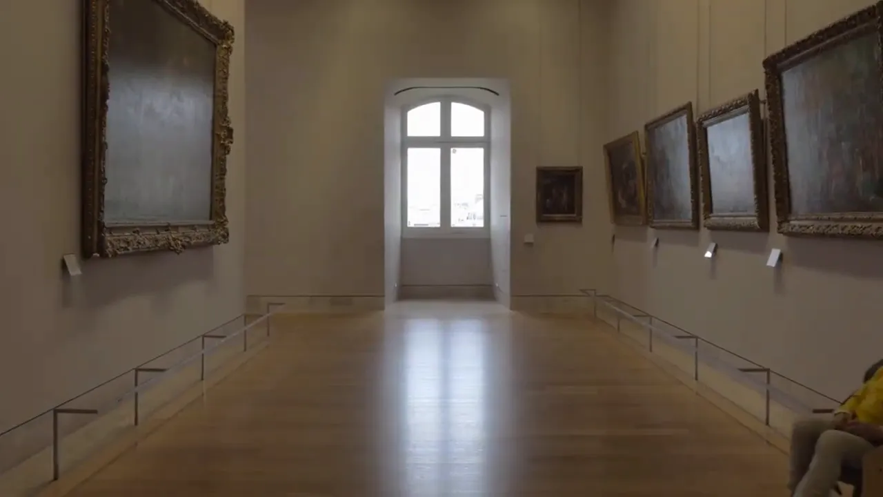 Interior view of an art gallery with framed paintings along the walls and a bright window at the far end