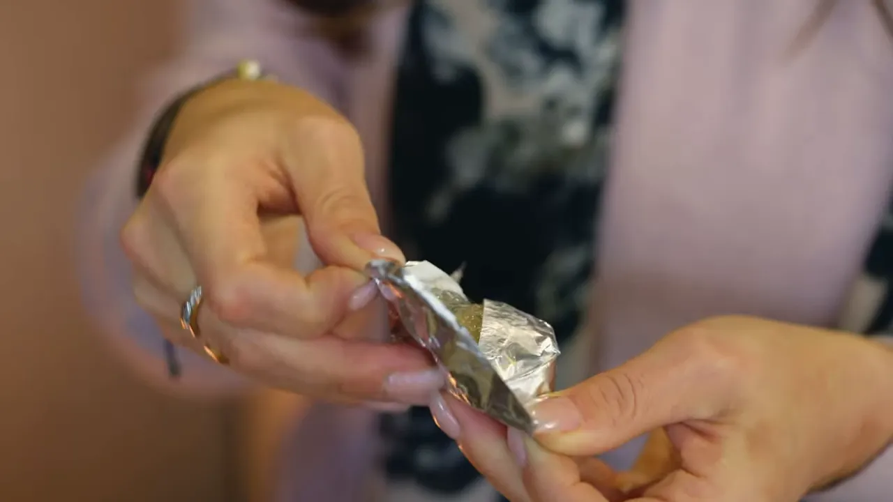 Close-up of hands unwrapping foil to show a piece of chestnut helva