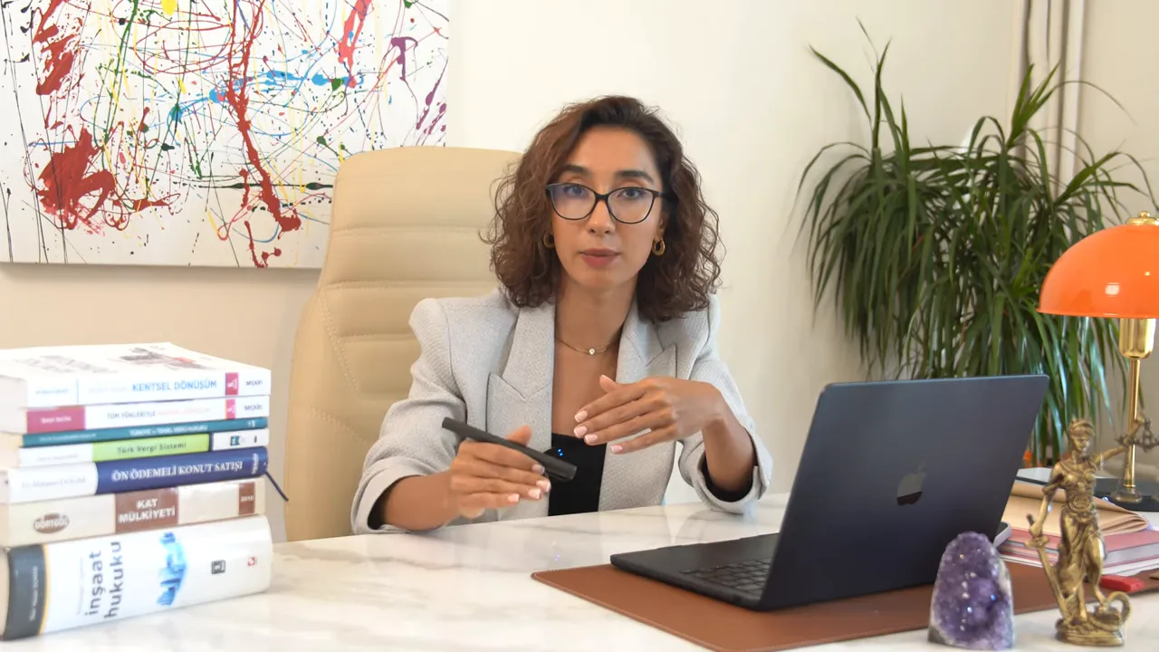 Ofiste masada oturan konuşmacı, elinde kalemle projeyi işaret eder gibi konuşuyor; dizüstü bilgisayar ve hukuk kitapları görünür.