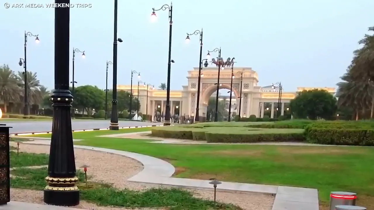 Gated palace entrance with arch, manicured lawns and decorative lamp posts in daylight.