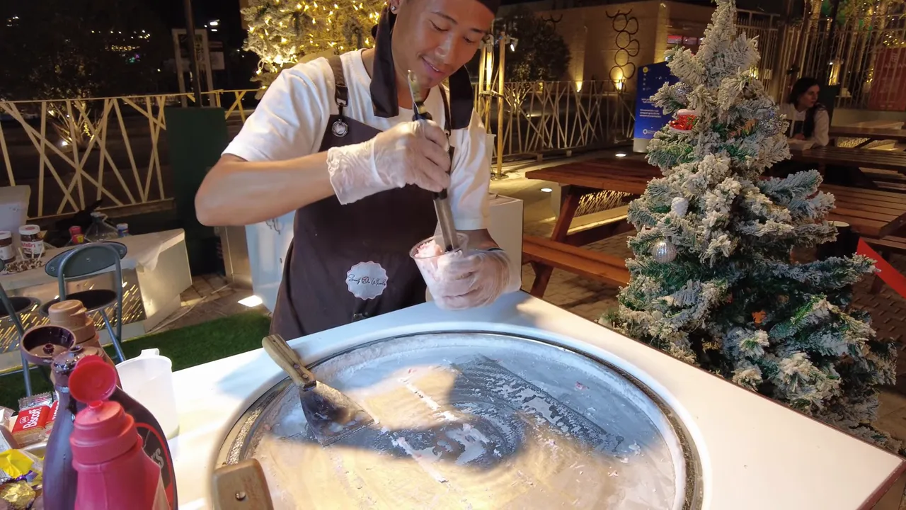 Close view of a vendor preparing rolled ice cream on a chilled pan and packing it into a cup with a snow‑flocked Christmas tree nearby