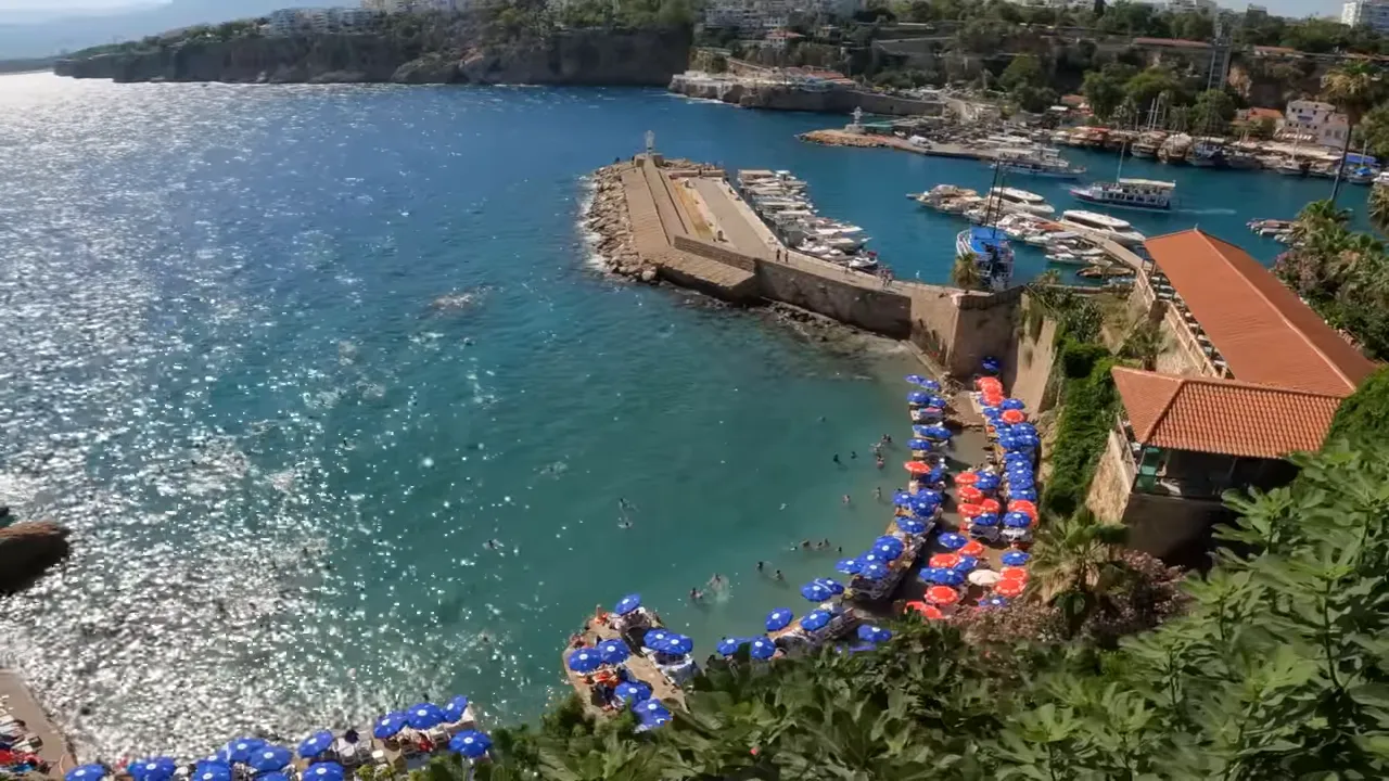 Mermerli Plajı kuşbakışı net görüntü; küçük koy, iskele, çok sayıda mavi ve kırmızı plaj şemsiyesi ve berrak turkuaz deniz.