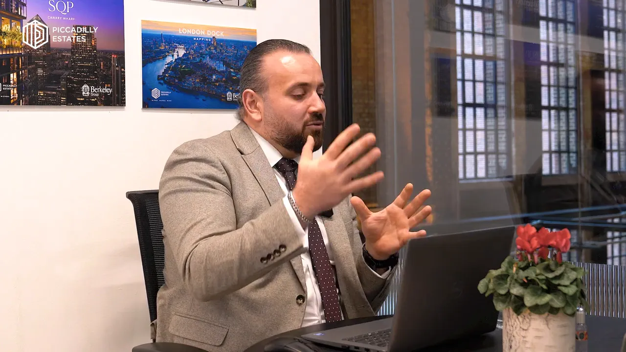 Diz&uuml;st&uuml; bilgisayar &ouml;n&uuml;nde el hareketleriyle konuşan takım elbiseli bir kişi; arka planda proje g&ouml;rselleri ve ofis manzarası.