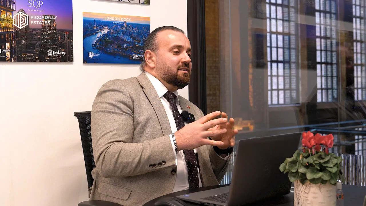 Ofis masasında diz&uuml;st&uuml; bilgisayar ve emlak posterleri &ouml;n&uuml;nde konuşan uzmanın geniş a&ccedil;ı g&ouml;r&uuml;nt&uuml;s&uuml;