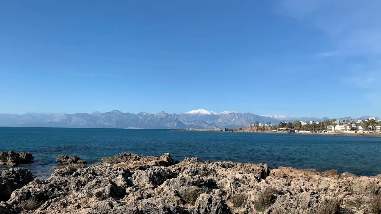 Konyaalti coastline with rocky foreground, blue sea and snow‑capped mountains on the horizon