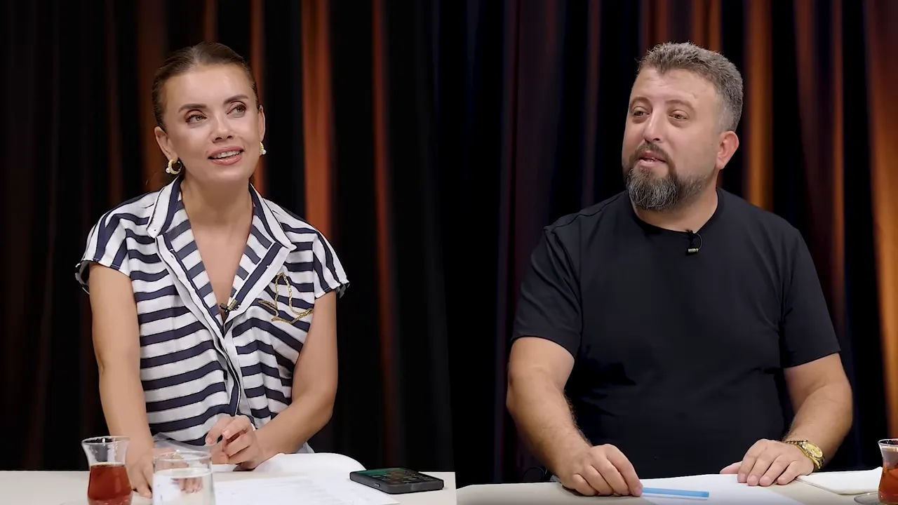 İki sunucunun masanın arkasında oturup konuştuğu geniş plan, kadın ve erkek sunucu birlikte